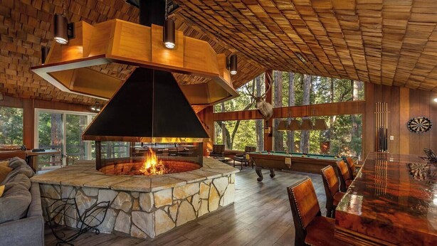A spacious indoor area with a stone fireplace, wooden ceiling, and large windows offering a view of the outdoors. - Camp Nelson Chalet + Bunkhouse | Hot Tub | Game Room (Springville)