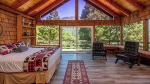 A log cabin bedroom with a large bed, two chairs, a desk, and a view of the forest. - Camp Nelson Chalet + Bunkhouse | Hot Tub | Game Room (Springville)
