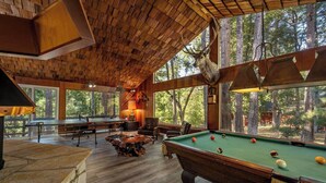 A pool table with a rustic wooden ceiling and large windows overlooking a forest. - Camp Nelson Chalet + Bunkhouse | Hot Tub | Game Room (Springville)