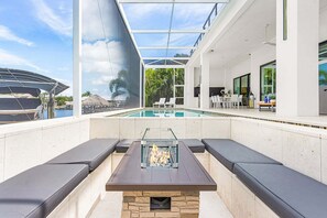 A modern outdoor pool area with a glass-enclosed fire pit, seating, and a view of the pool. - Luxury Gulf Access Villa with Pool, Spa & Waterfront Fun Escape - Villa Metropolis - Roelens (Cape Coral)