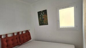 Ein Einzelzimmer mit einem weißen Bett, einem roten Kissen an der Kopfteil, einer Deckenleuchte, einem Fenster mit Jalousien und einem kleinen Wandbild.