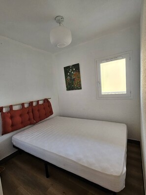 A single-bed room with a white mattress, a headboard with red cushions, a ceiling light fixture, a window with blinds, and a small wall-mounted artwork. - Lovely 2-bedroom apartment near Richelieu beach, Cap d'Agde (LE CAP D AGDE)