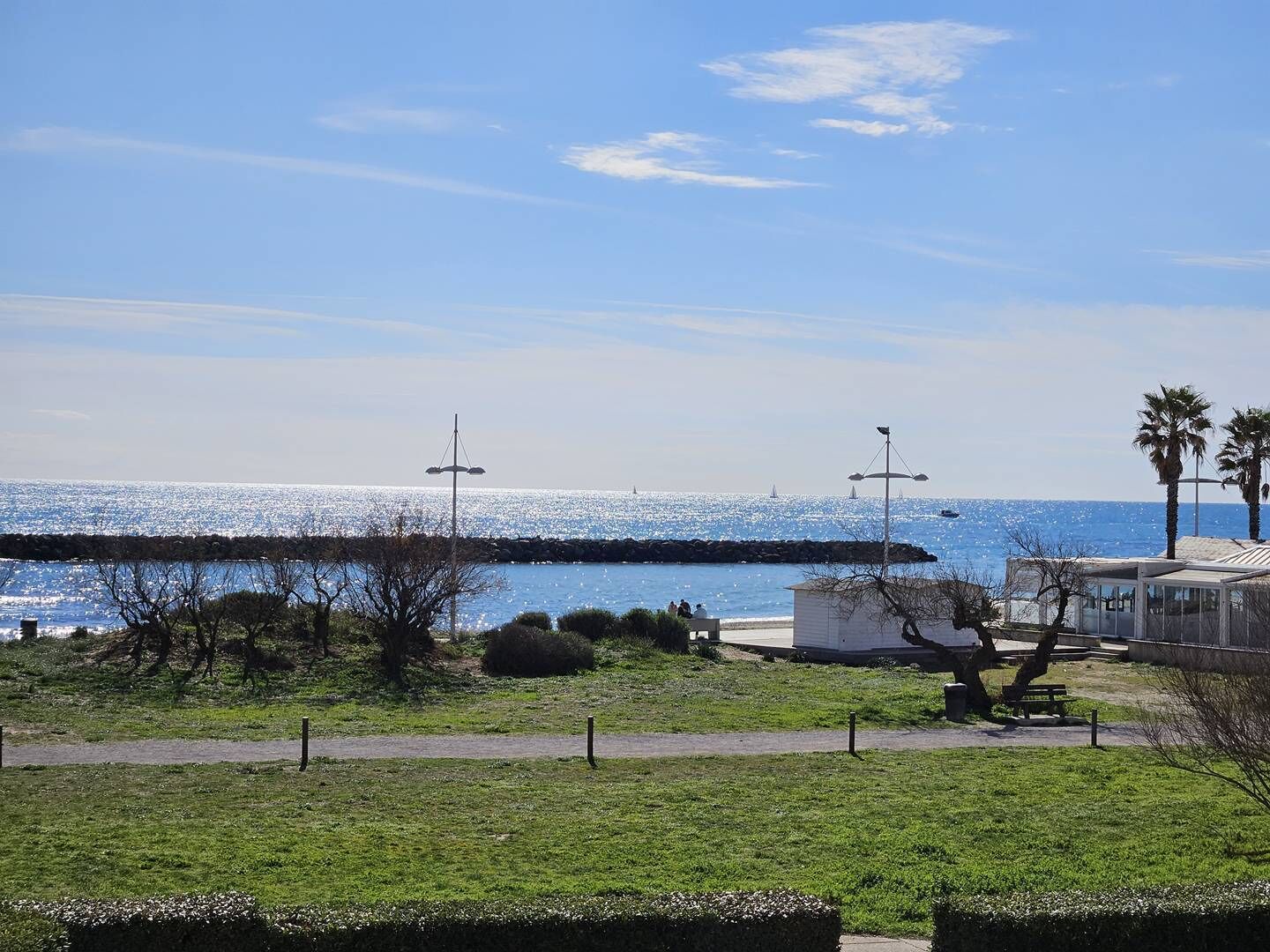 Vista costeira com céu claro, um molhe rochoso e uma área gramada com um banco.