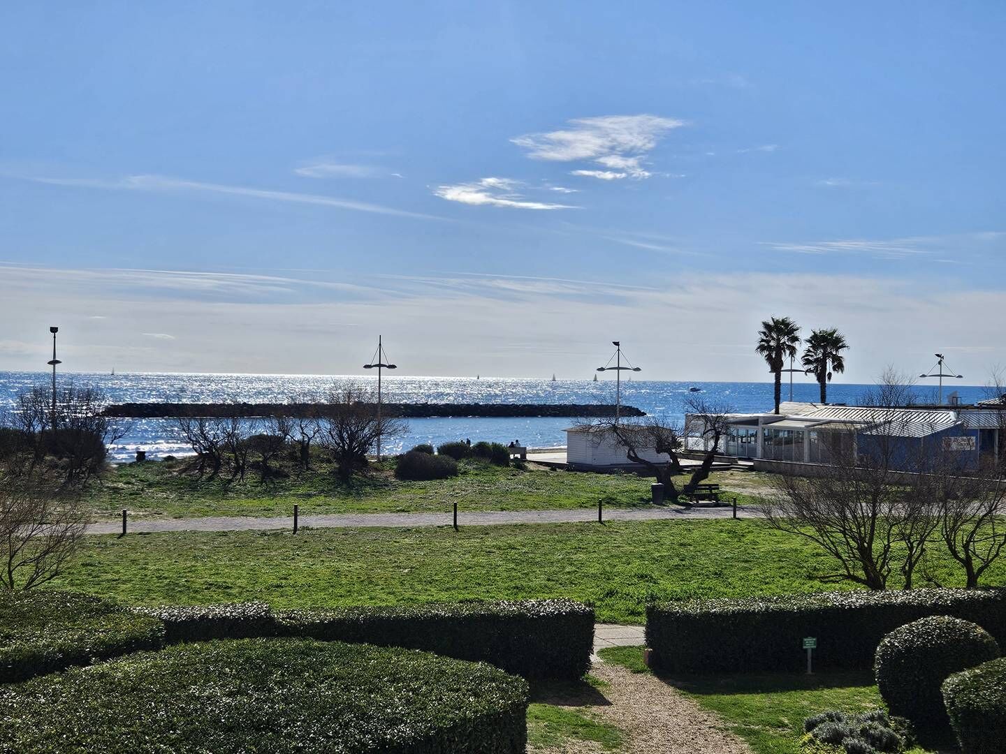 Blick auf die Küste mit blauem Himmel, einer sich ins Wasser erstreckenden Pier und einer Rasenfläche mit geschnittenen Büschen.