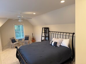 Um quarto com uma cama, um ventilador de teto, um violão, uma cadeira e uma estante de livros.