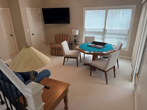 A living room with a TV, a plaid armchair, a table with a board game, and a window with blinds.