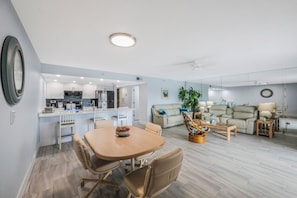 Eine moderne Küche mit weißen Schränken, ein Essbereich mit Holztisch und -stühlen sowie ein Wohnzimmer mit Sofa und Couchtisch.