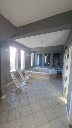 Een ruime kamer met een groot bad, een hoek in het zonlicht met twee witte stoelen, en uitzicht op de stad door een raam.