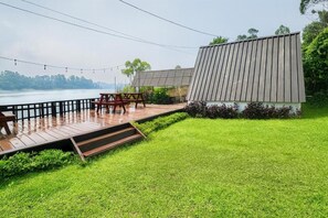 A deck with a table and chairs overlooking a body of water, a building with a sloped roof, and a grassy area. - Cozzy Villa Tb (BANDUNG)