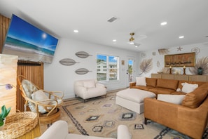 Ein Wohnzimmer mit Strandkunst an der Wand, einem Korbstuhl, einem weißen Sofa, einem braunen Ecksofa und einem gemusterten Teppich.