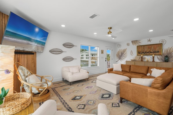 A living room with a beach-themed wall art, a wicker chair, a white sofa, a brown sectional sofa, and a patterned rug. - Livin' Largo | Dog-Friendly | Fenced-In Backyard | My Beach Getaways (Gulf Breeze)