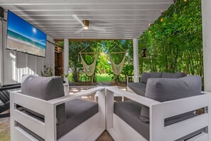 Un espacio moderno al aire libre con mobiliario blanco y vistas a un jardín exuberante y una playa.