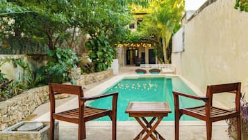 A pool area with two wooden chairs and a small table, surrounded by lush greenery and a stone wall.