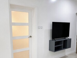 Ein Zimmer mit einer weißen Tür, Fensterflächen mit gebürstetem Glas, einem montierten Fernseher auf einem grauen Regal und einem Lichtschalter an der Wand.