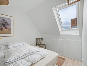 A bedroom with a sloped ceiling, a bed with white bedding, a chair, a framed picture, and a window with a view of a building. - "Swarthofthi" - 90m from the sea by Interhome (Assens)