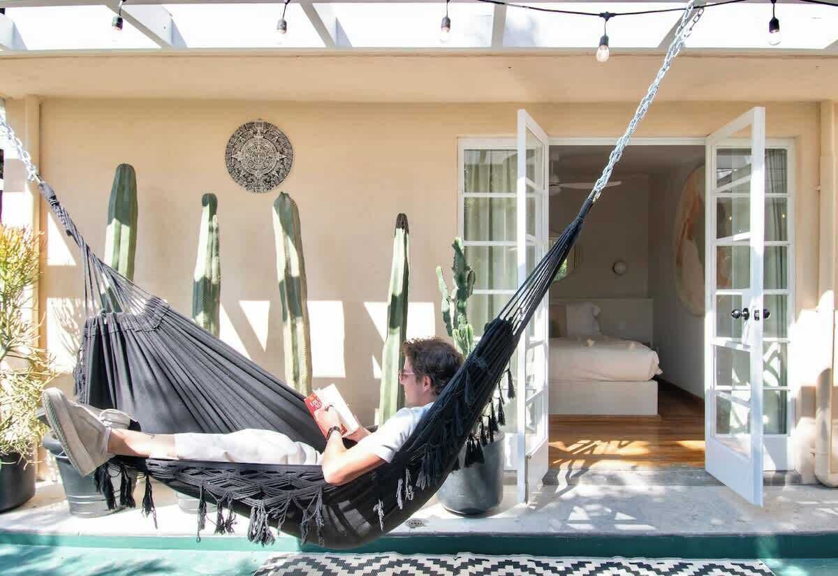 Uma pessoa relaxando em uma rede em uma varanda com vista para um jardim de cactos e uma porta que dá para um cômodo interno.