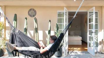 Uma pessoa relaxando em uma rede em uma varanda com vista para um jardim de cactos e uma porta que dá para um cômodo interno.