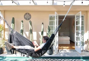 Eine Person entspannt sich in einer Hängematte auf einem Balkon mit Blick auf einen Kakteengarten und einer Tür, die zu einem Innenraum führt.