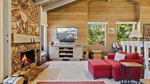 Un salon chaleureux avec une cheminée en pierre, un téléviseur, un canapé rouge et une vue sur la verdure à l’extérieur.