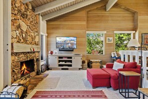 Ein gemütliches Wohnzimmer mit Kamin aus Stein, einem Fernseher, einem roten Sofa und Blick auf Grünfläche.