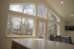A kitchen with large windows, a dining area with chairs, and a TV mounted on the wall. - Welcome to Sky Ranch, a spacious, warm newly built home ready to welcome you. (Talkeetna)