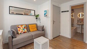 Una sala de estar moderna con un sofá gris, una mesita blanca, un espejo y una puerta que da a un baño.