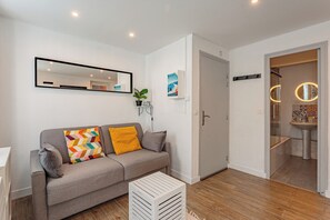 Ein modernes Wohnzimmer mit einem grauen Sofa, einem weißen Beistelltisch, einem Spiegel und einer Tür zu einem Badezimmer.