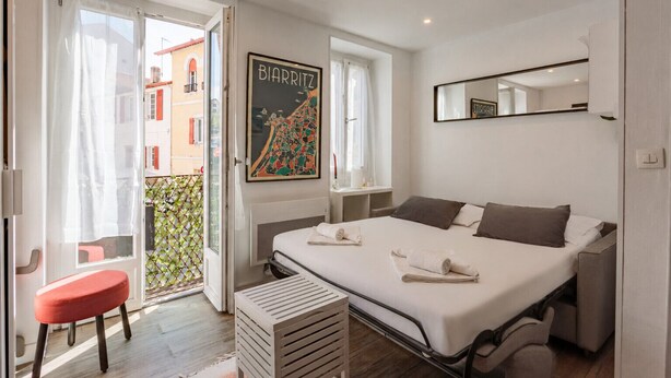 Un dormitorio con una cama, un taburete rojo, una ventana con vistas y un cuadro en la pared.
