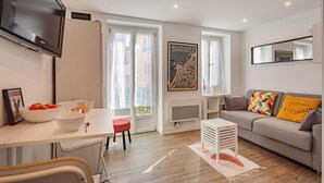 Una sala moderna con un sofá, una mesa de comedor con sillas, un televisor montado en la pared y una ventana con cortinas.