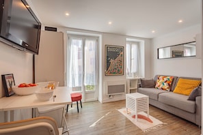 Ein modernes Wohnzimmer mit einer Couch, einem Esstisch mit Stühlen, einem an der Wand montierten Fernseher und einem Fenster mit Vorhängen.