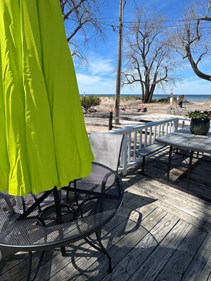 Eine Holzterrasse mit einem Metalltisch und -stühlen, einem leuchtend grünen Sonnenschirm und Blick auf Strand und Meer.