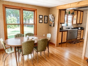 A dining area with a wooden table and chairs, a kitchen with wooden cabinets, and a view of the outdoors through large windows.
