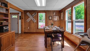 A dining area with a wooden table set for four, a bookshelf, and a view through large windows. - New Listing! 4 Bedroom Home 2 mins from the Lake-Boating-Fishing-Fire Pit (Trout Lake)