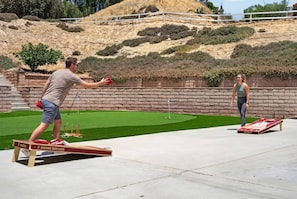 Una persona juega al shuffleboard sobre una superficie de concreto, con un césped verde y un muro de ladrillo al fondo.