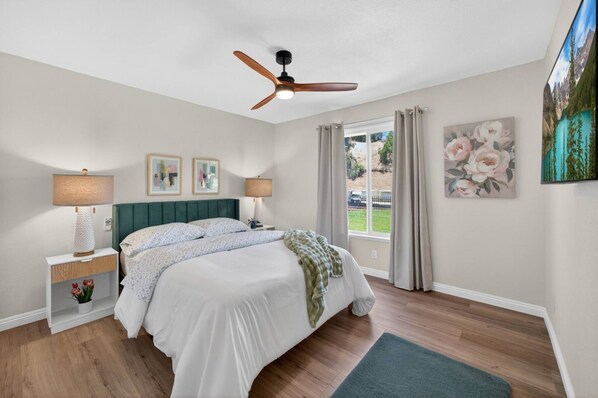 A bedroom with a bed, two bedside tables, a ceiling fan, a window with curtains, and wall art.
