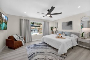 A modern bedroom with a bed, a chair, a ceiling fan, a window with curtains, and a view of the outdoors.