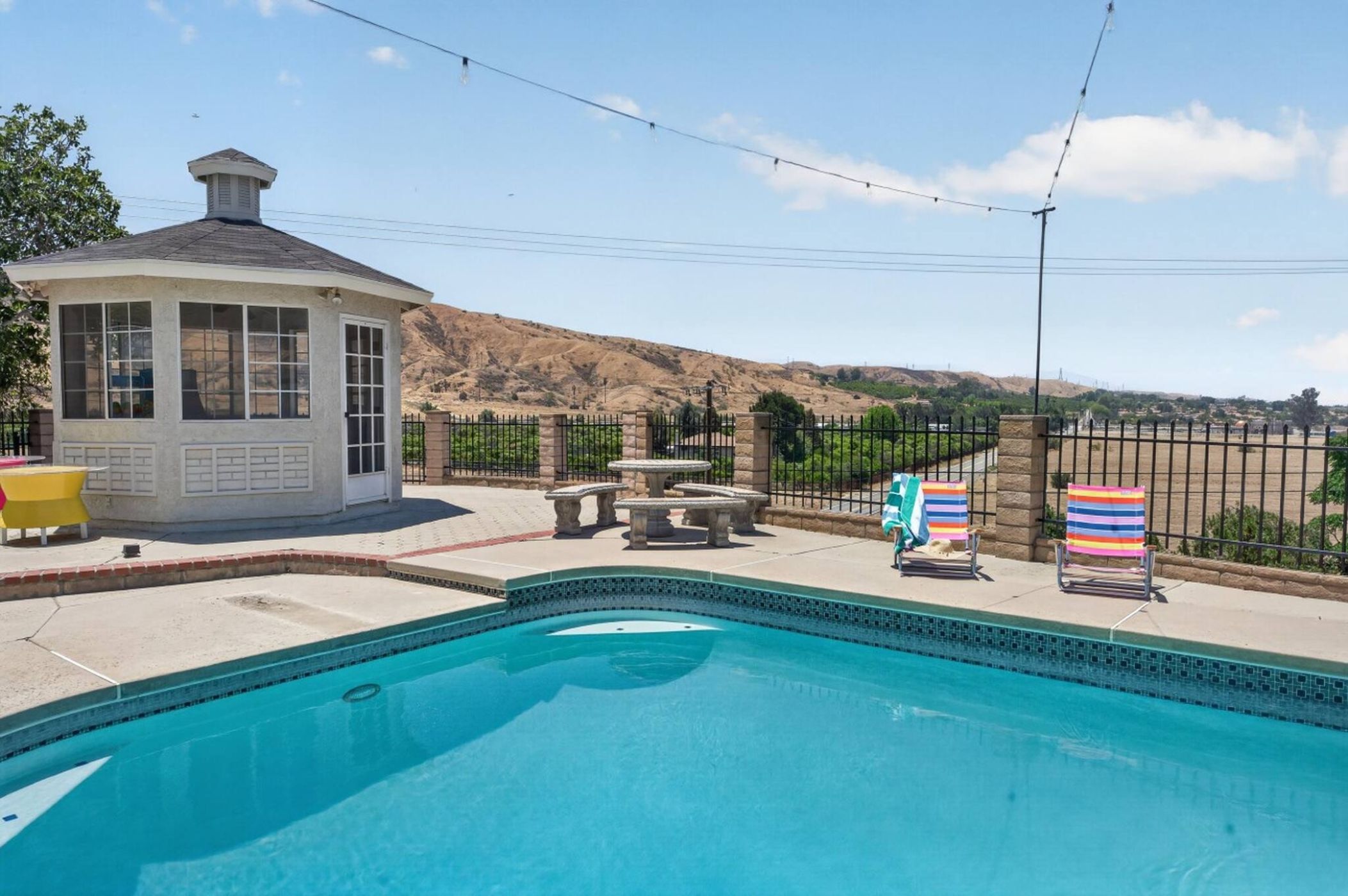 A swimming pool with a fenced area, a small building with windows, and a hilly landscape in the background.
