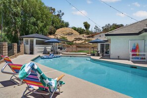 A swimming pool with lounge chairs and umbrellas, surrounded by a landscaped area with trees and hills in the background.