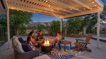 A covered outdoor seating area with a fire pit, lounge chairs, and string lights.