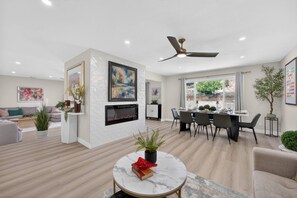 A modern living room with a fireplace, dining area, and a ceiling fan. - Executive Corporate Stay | 4BR/4BA Pool-Spa & Golf (Redlands)