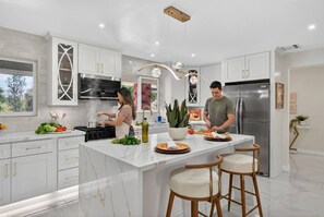 A modern kitchen with white cabinetry, a central island with stools, a stainless steel refrigerator, and a large window. - Executive Corporate Stay | 4BR/4BA Pool-Spa & Golf (Redlands)