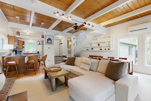 A spacious living area with a sofa, coffee table, and chairs, a kitchen with bar stools, and a staircase leading to another level.
