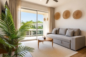 Ein modernes Wohnzimmer mit einem grauen Sofa, einem runden Couchtisch und einem großen Fenster mit Blick auf Grünflächen.