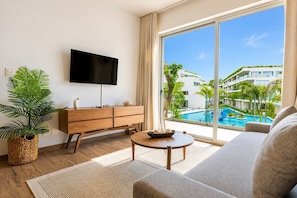 Ein modernes Wohnzimmer mit Flachbildfernseher, Holzmöbeln und einem großen Fenster mit Blick auf einen Pool und Gebäude.