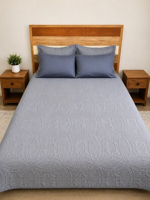 Ein Bett mit gemustertem Überwurf, zwei Kissen, einem Holz-Kopfteil und zwei Nachttischen, auf einem davon steht eine kleine Pflanze.