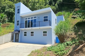 A modern two-story building with a balcony, a concrete driveway, and surrounding greenery. - Your Caribbean Escape – Lovely 2-Bedroom Apartment in Cochrane, Dominica (Cochrane)