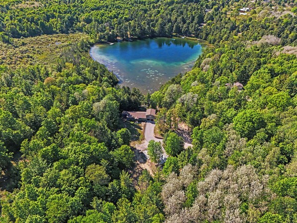 An aerial view of a lake surrounded by dense forest, with a small building complex visible in the foreground.