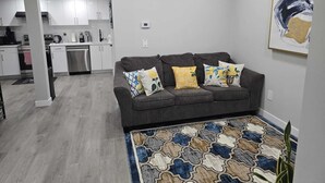 A modern living room with a grey sofa, patterned rug, and a kitchen in the background. - Brand New Luxury Basement (Calgary)