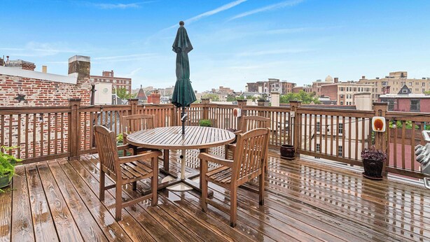 A wooden deck with a round table and chairs, a green umbrella, and potted plants. - Sojourn on Swann | Chic 2BD with Rooftop Deck (Washington DC)