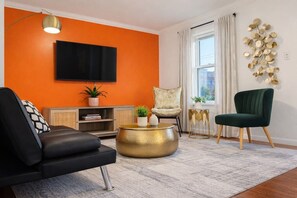 Ein Wohnzimmer mit einer schwarzen Couch, einem grünen Sessel, einem runden Couchtisch aus Gold, einem Flachbildfernseher und einem Fenster mit Vorhängen.